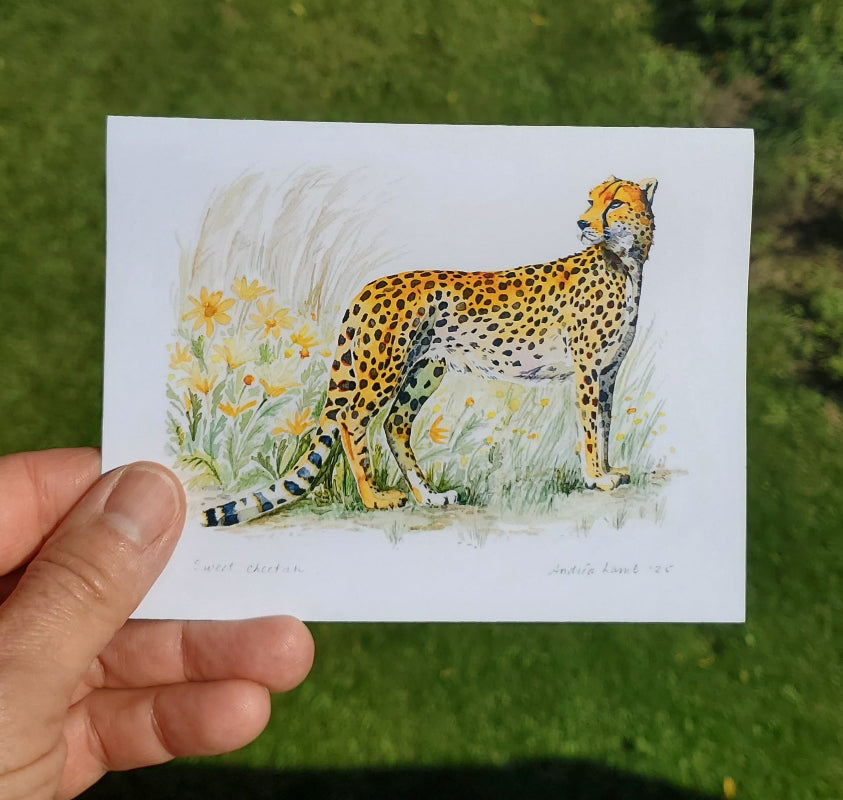 A hand holds a small art print depicting a cheetah standing among yellow flowers on a white background, with green grass visible behind it.
