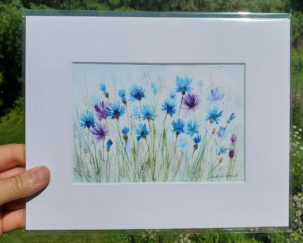 Matted artwork of blue and purple flowers held by a hand with a natural background.