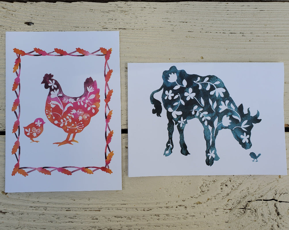 Two decorative art cards displayed side by side on a rustic, light-colored wooden surface. The left print features a stylized red and orange chicken and chick with a floral leaf border.