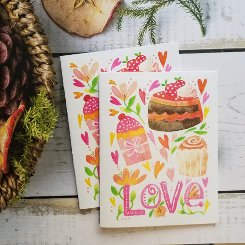 Two colorful Valentine cards with illustrations of cakes and flowers on a wooden surface.
