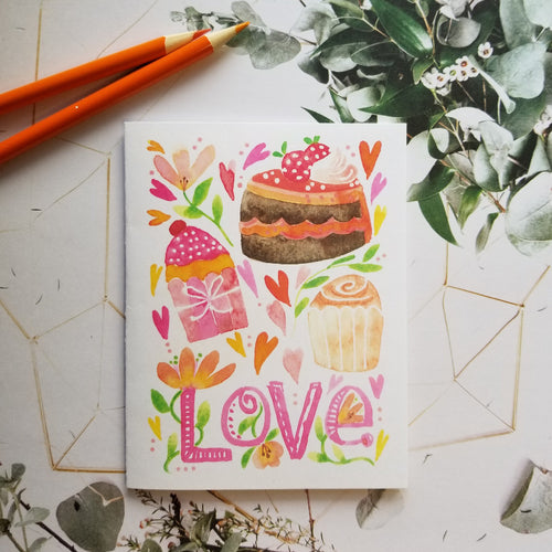 Valentine card with colorful illustrations of cakes and flowers on a white surface with greenery.