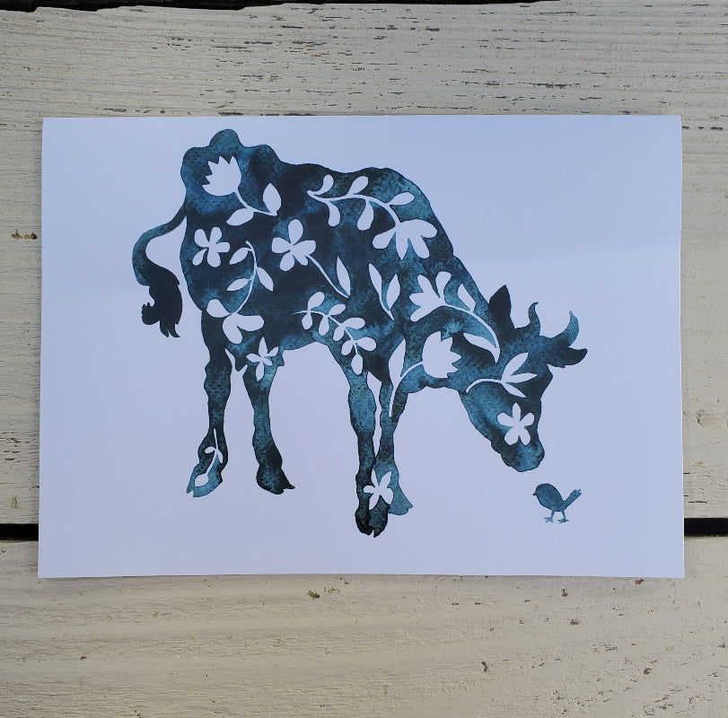 A fine art card featuring a stylized blue cow with white floral patterns and a small bird, displayed on a light wood surface.