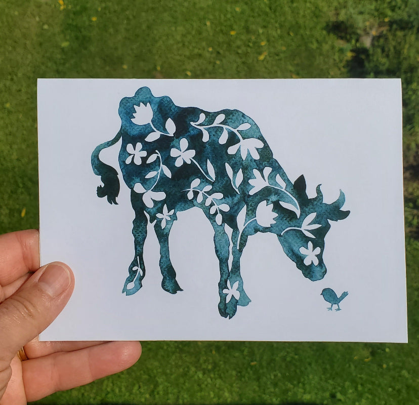 A hand holds a fine art card featuring a blue watercolor-style cow decorated with white floral motifs, set against a blurred green grassy background.
