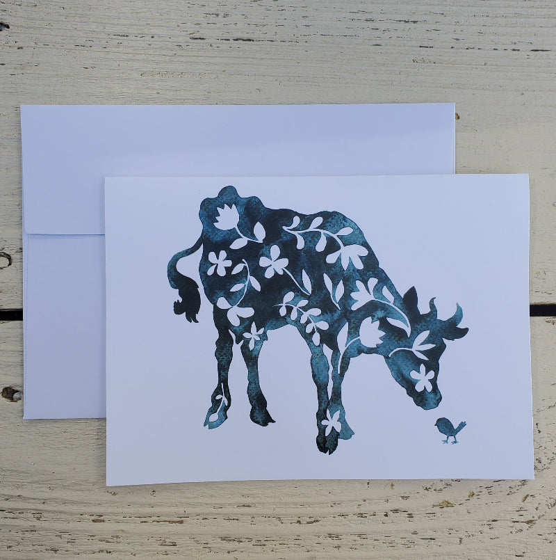 A white greeting card featuring a blue watercolor-style cow filled with white floral patterns, shown with a matching white envelope on a light wood surface.