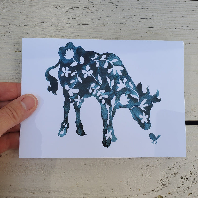 A hand holds a white fine art card featuring a stylized cow in dark blue watercolor with white floral details and a small bird, set against a light wood background.