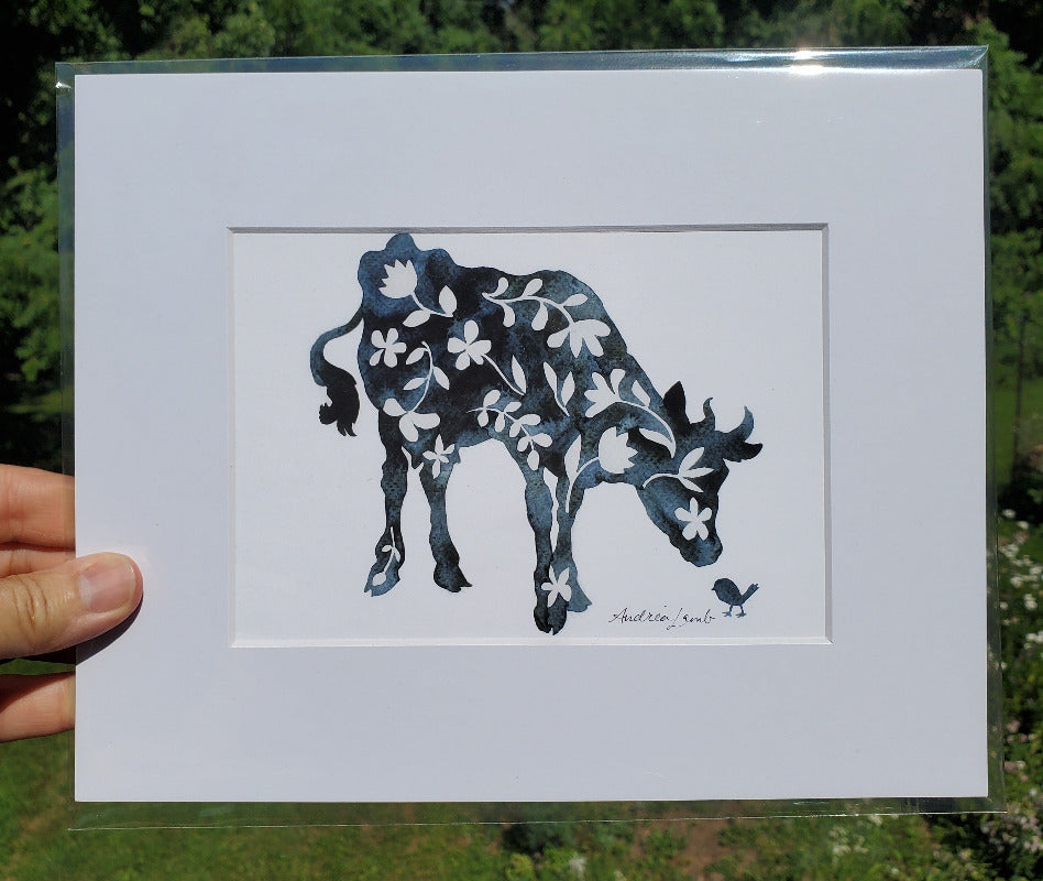 Matted artwork of a stylized cow with floral patterns held in front of a natural background.
