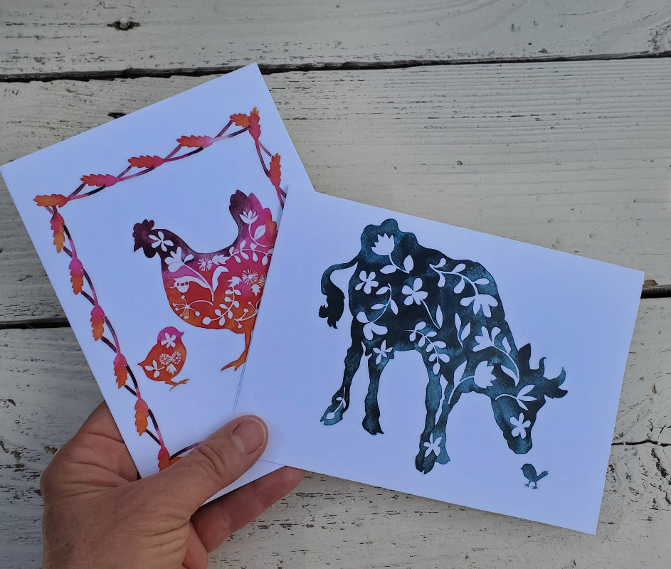 A hand holds two white art cards featuring stylized farm animals with floral patterns, set against a rustic white wooden plank background. One card shows a red and orange chicken and chick, and the other shows a dark blue cow with white floral motifs.