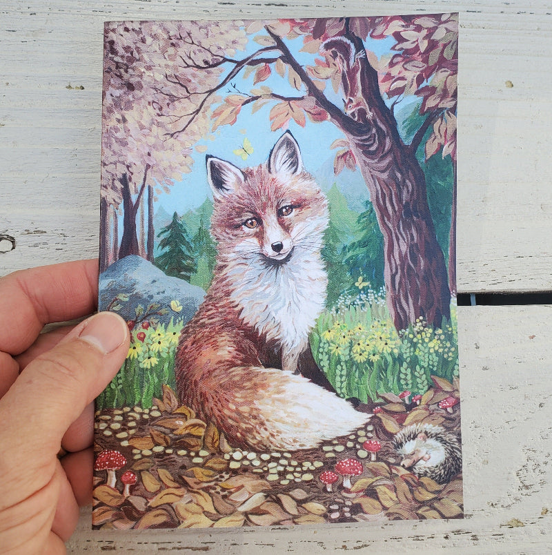 A hand holds a rectangular fine art card featuring a painted fox in a forest scene with autumn trees, flowers, and leaves. The print is photographed against a distressed white wooden surface.