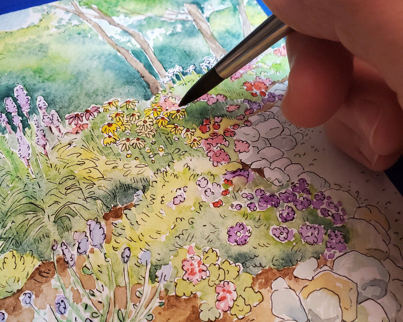 Hand holding a paintbrush over a watercolor painting of a garden scene with flowers and rocks.