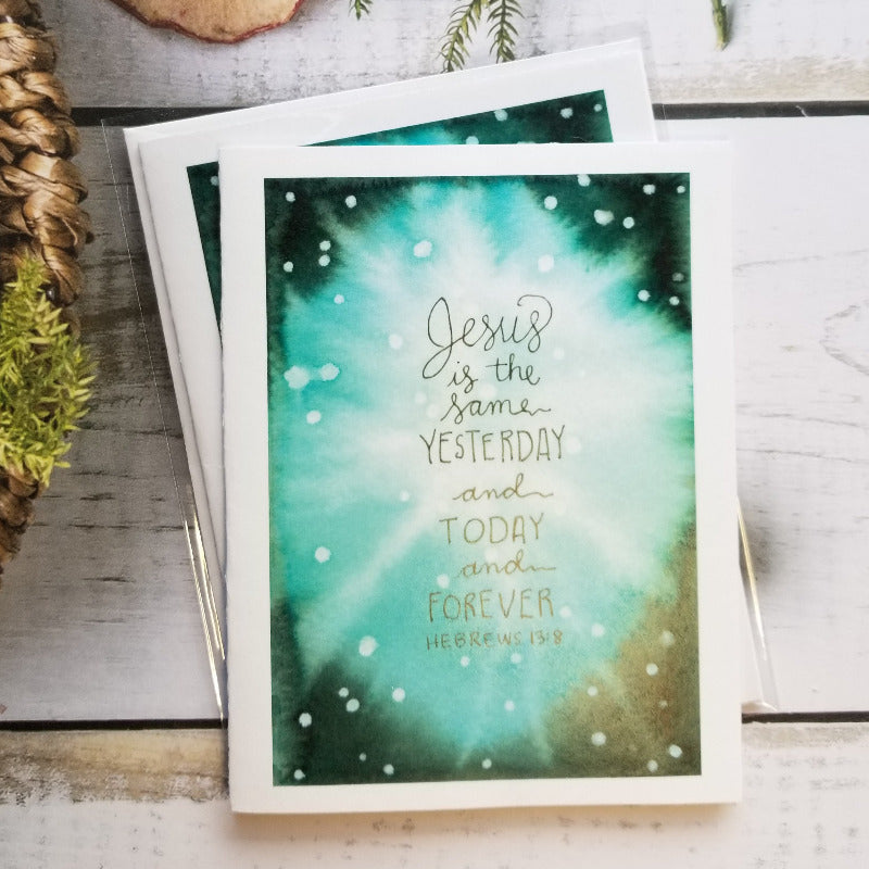 Card with 'Jesus is the same yesterday and today and forever' text on a decorative surface with pinecones and branches.
