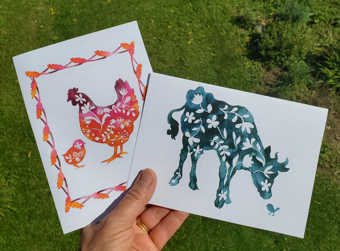 A hand holds two printed art cards outdoors over a green grassy background. One card shows a colorful floral chicken and chick with orange and pink tones, and the other shows a dark teal floral cow with white floral details.