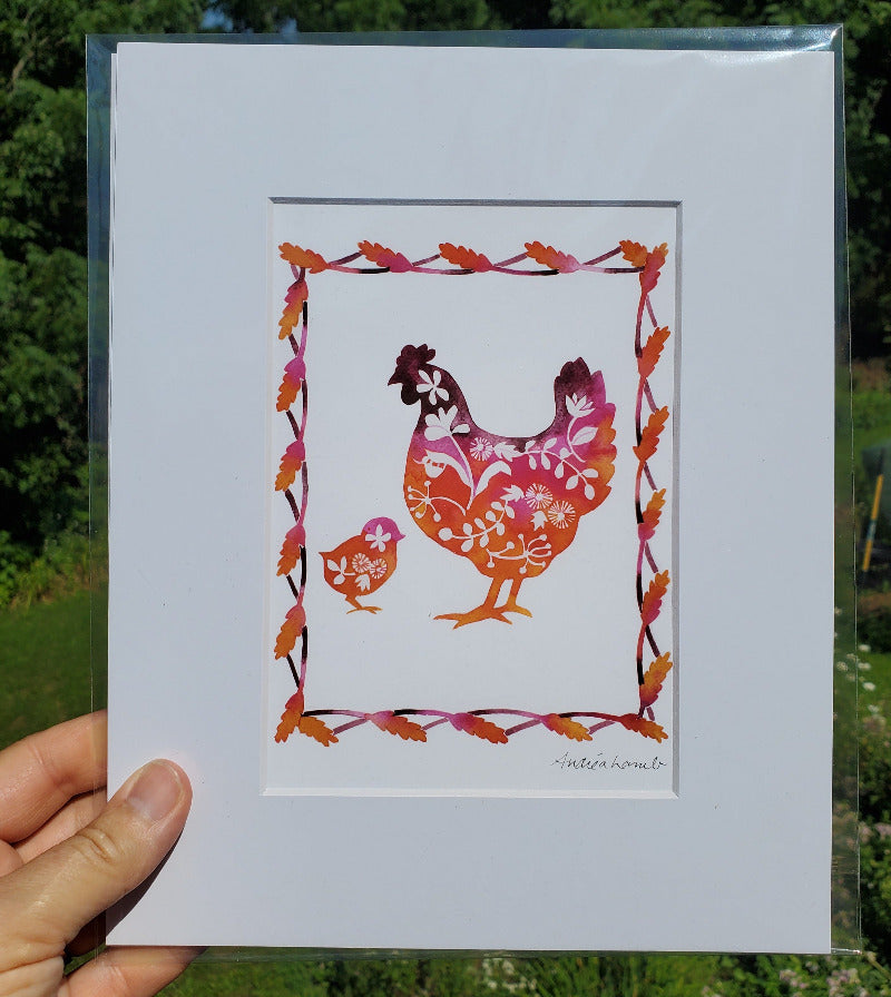 Decorative print of a rooster and chick with floral patterns held against a natural background.