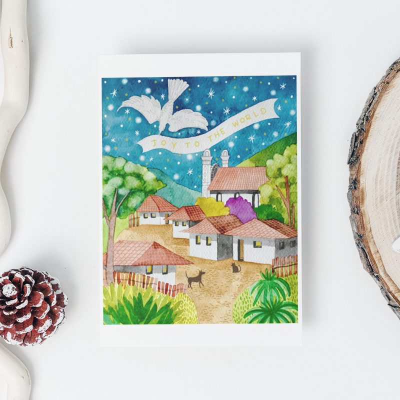 Framed artwork of a snowy village scene with pine cones and wood slice on a white background