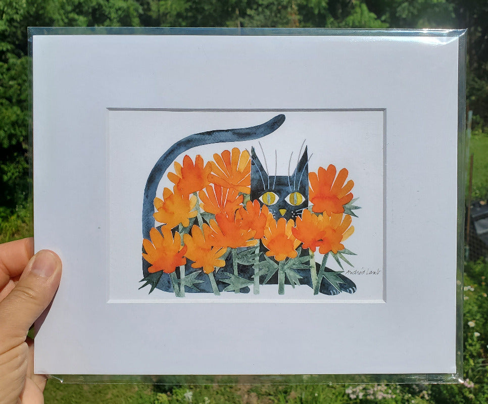 Matted artwork of a cat among orange flowers held by a person outdoors.