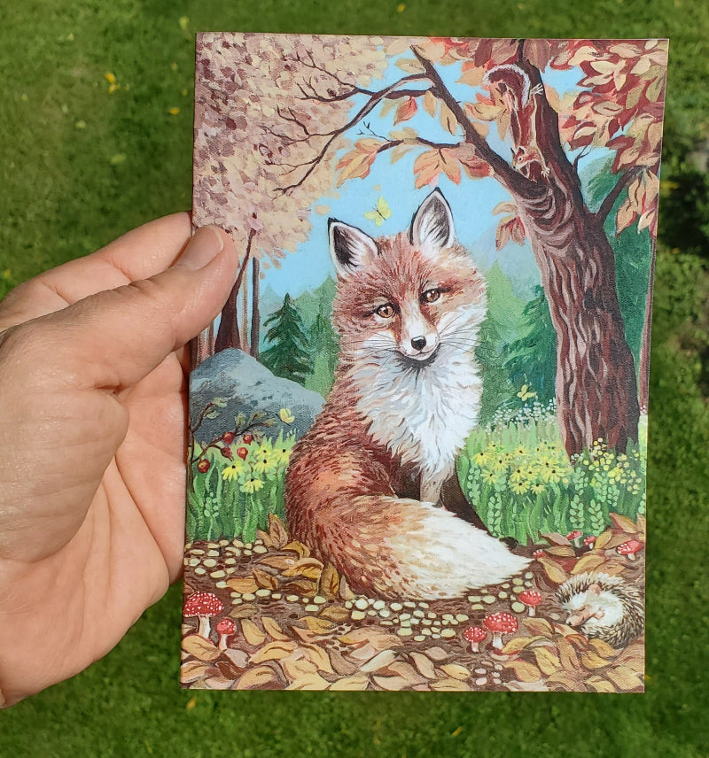 A hand holds a fine art card featuring a painted fox in an autumn forest, with colorful foliage, flowers, and trees. The background is real grass outdoors.
