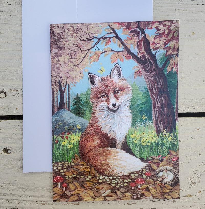 A fine art card featuring a painted fox in an autumn forest, placed on a light wood surface with a white envelope behind it.