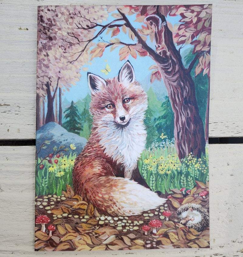 A vertical fine art card depicting a detailed illustration of a red fox in a colorful autumn forest, placed flat on a light-colored wooden surface.