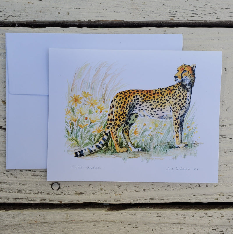 A fine art card featuring a painted cheetah standing in grass with yellow flowers, placed in front of a white envelope on a weathered light wood surface.